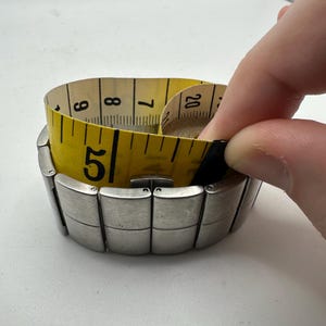 May include: A silver metal watch band with a yellow measuring tape wrapped around it. The tape measure reads 5, 6, 7, 8, and 20.