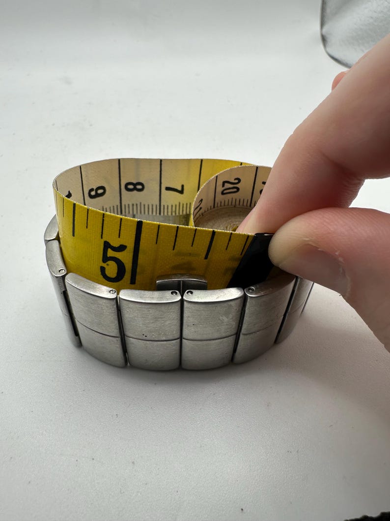 May include: A silver metal watch band with a yellow measuring tape wrapped around it. The tape measure reads 5, 6, 7, 8, and 20.