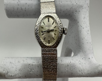 May include: A vintage Bulova wristwatch with a silver oval face and a textured silver band. The watch face displays the Bulova logo and the number 23. The band is a flexible, textured design.