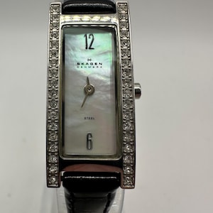 Op de afbeelding: Een zilveren Skagen Denmark horloge met een witte parelmoer wijzerplaat en een zwarte leren band. Het horloge heeft een rechthoekige wijzerplaat met een diamant bezel en een zilveren band. De wijzerplaat van het horloge heeft zilveren wijzers en Romeinse cijfers als uurmarkeringen.