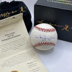Pode incluir: Uma bola de beisebol branca assinada por Duke Snider, um jogador de beisebol do Hall da Fama. A bola está em uma caixa preta com um rótulo branco que diz "Authentic Memorabilia". O rótulo também inclui o texto "We guarantee this Duke Snider HOF 80 Autographed Baseball to be a genuine hand-signed piece of memorabilia. Our seal of authenticity guarantees the integrity of this product. Any attempt to remove the seal will void the authenticity of this collectible. This item was signed in the presence of a Reggiejackson.com employee."