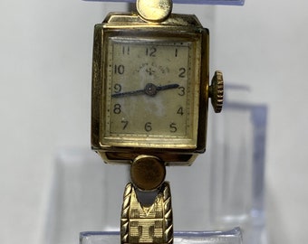 May include: A gold-toned Lady Elgin wristwatch with a square face and a gold-toned metal band. The watch face has black Roman numeral hour markers and black hands.