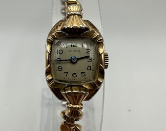 May include: A gold-toned wristwatch with a square face and a gold-toned metal band. The watch face has black Roman numeral hour markers and a white background. The watch face also has the word "Buren" printed on it.