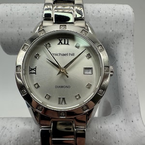 May include: A silver-toned Michael Hill wristwatch with a round face. The watch features diamond accents around the bezel and on the face, with Roman numeral hour markers. The watch band is a matching silver color.