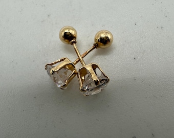 May include: Two pairs of gold-coloured stud earrings. One pair features a small, round gold ball, and the other has a clear, faceted gemstone set in a gold bezel. The earrings are displayed on a light-coloured surface.