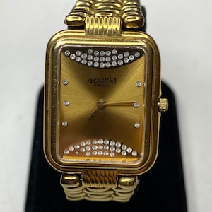 May include: Gold-toned Heloisa Swiss watch with a rectangular face and a gold band. The watch face is gold with a sunburst pattern and two rows of small, clear crystals. The watch has the brand name "HELOISA SWISS" printed on the face.