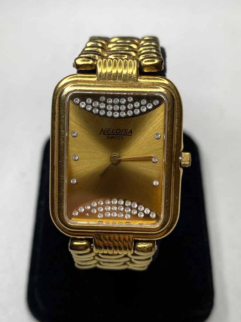 May include: Gold-toned Heloisa Swiss watch with a rectangular face and a gold band. The watch face is gold with a sunburst pattern and two rows of small, clear crystals. The watch has the brand name "HELOISA SWISS" printed on the face.