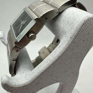 May include: A silver wristwatch with a square face and a black dial. The watch has a silver metal band with a brushed finish.