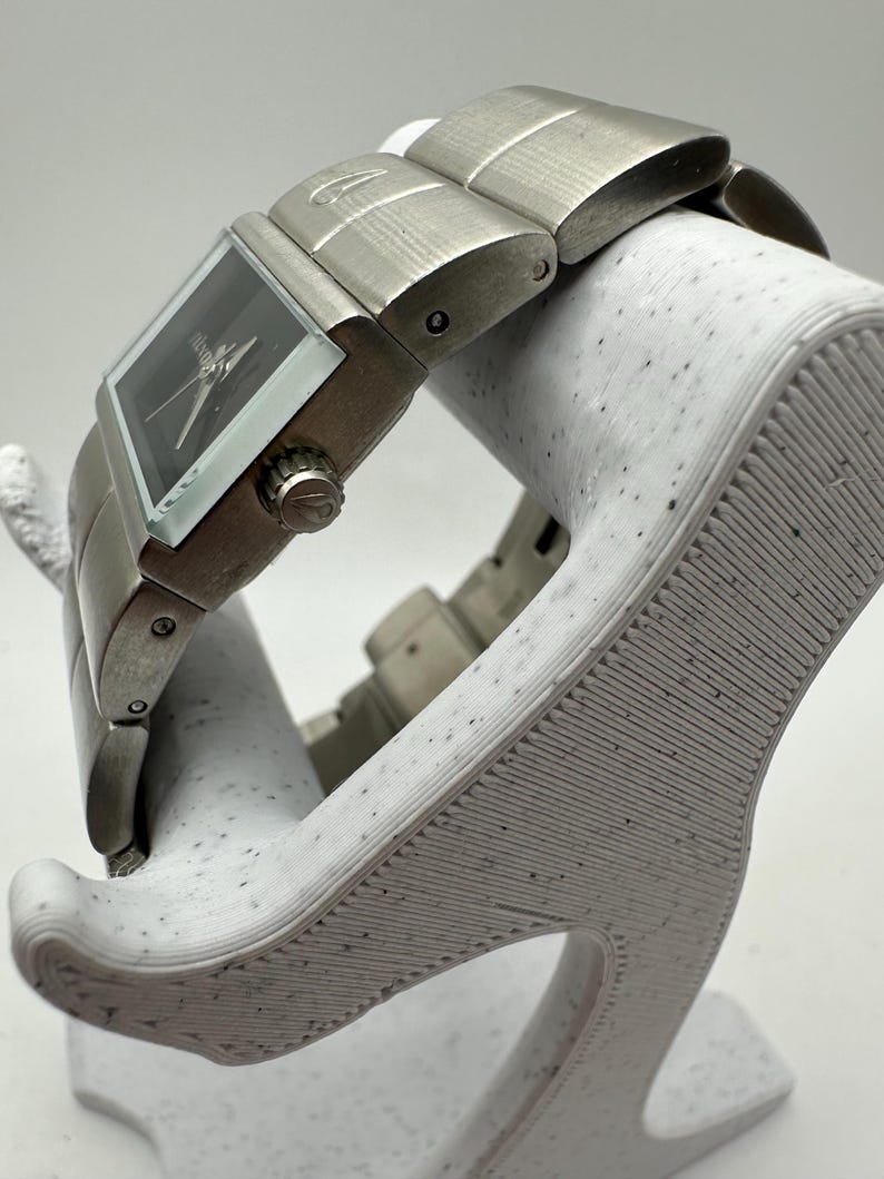 May include: A silver wristwatch with a square face and a black dial. The watch has a silver metal band with a brushed finish.