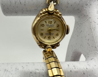 May include: A gold-toned Delroy 17 Jewels wristwatch with a white face and black numerals. The watch has a gold-toned metal band.