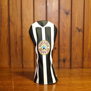 May include: A black and white striped golf club head cover with a Newcastle Brown Ale logo. The cover has a V-neck design and is made of a synthetic material. The background is a wooden surface.