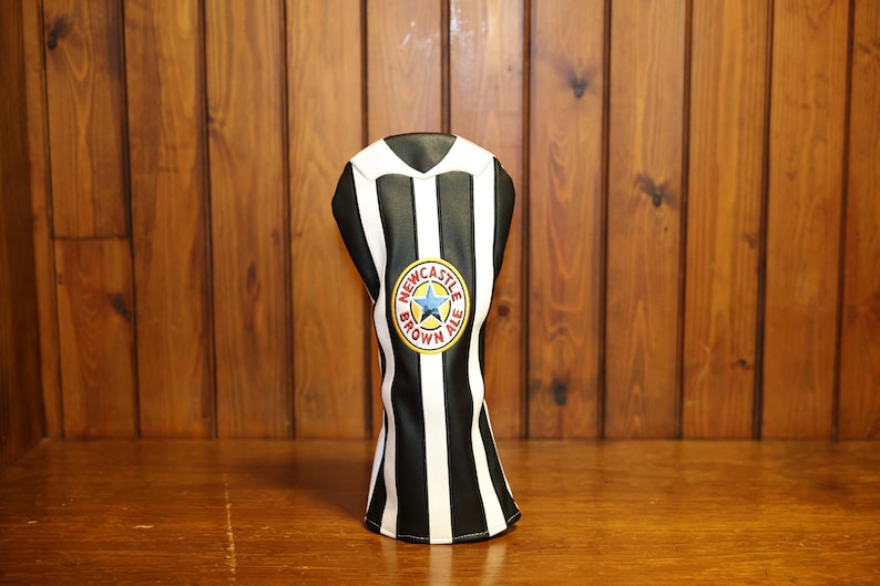 May include: A black and white striped golf club head cover with a Newcastle Brown Ale logo. The cover has a V-neck design and is made of a synthetic material. The background is a wooden surface.
