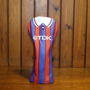 Op de afbeelding: Een golfclub headcover ontworpen als een sportshirt met verticale rode en blauwe strepen. De hoes heeft een witte kraag met twee knopen en de witte tekst TDK in het midden gedrukt. Het materiaal ziet er glad en synthetisch uit.