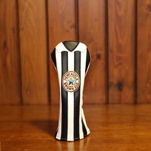 May include: A golf club head cover with a black and white striped design, resembling a soccer jersey. The cover features a circular emblem with text and a star, set against a wooden background.