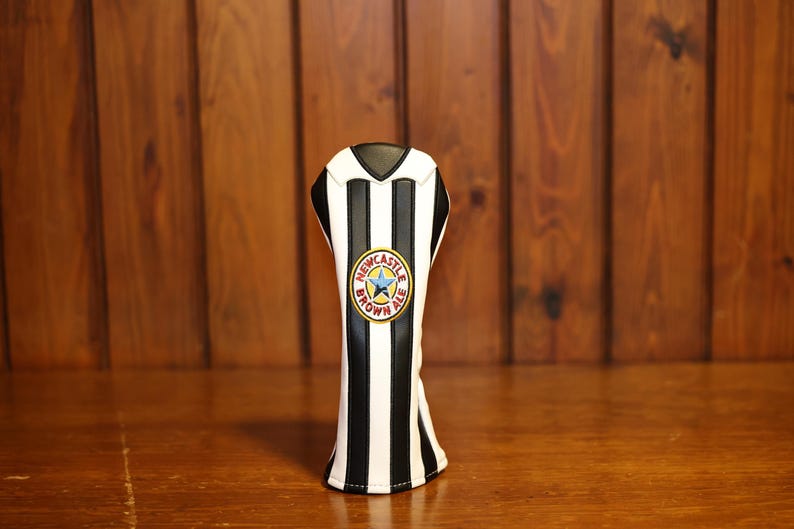 May include: A golf club head cover with a black and white striped design, resembling a soccer jersey. The cover features a circular emblem with text and a star, set against a wooden background.