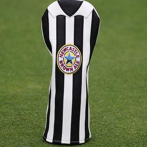 May include: A golf club head cover with a black and white striped design. The cover features a circular emblem with a blue star and the words "NEWCASTLE BROWN ALE" in a yellow circle. The cover is set against a green background.