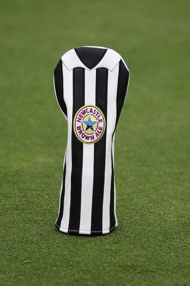 May include: A golf club head cover with a black and white striped design. The cover features a circular emblem with a blue star and the words "NEWCASTLE BROWN ALE" in a yellow circle. The cover is set against a green background.