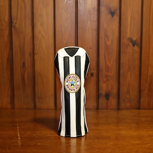 Peut inclure: Une housse de club de golf &agrave; rayures noires et blanches, orn&eacute;e d'un logo Newcastle Brown Ale. Le couvre-chef a un dessus noir et est pr&eacute;sent&eacute; sur un fond en bois.