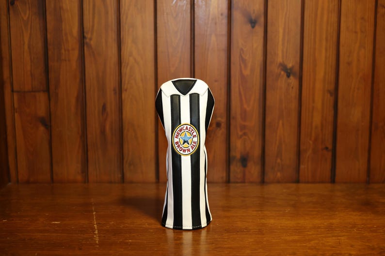 Peut inclure: Une housse de club de golf &agrave; rayures noires et blanches, orn&eacute;e d'un logo Newcastle Brown Ale. Le couvre-chef a un dessus noir et est pr&eacute;sent&eacute; sur un fond en bois.