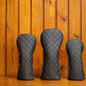 May include: Three black golf club head covers of varying sizes, each featuring a quilted diamond pattern and a small circular logo at the base. The covers are constructed with a smooth, matte finish and are arranged in a row from smallest to largest.