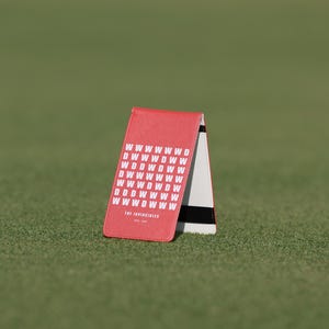 May include: A red and white golf scorecard holder with the text "WWWWWWWD DWWDWW WDDWDWW DWWWWWW WWWDWDW DDDDWWW WWWDWWW" and "THE INVINCIBLES" on the front. The scorecard holder is on a green surface.
