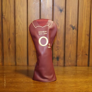 Peut inclure: Une housse de club de golf marron avec des lettres et des accents dorés. La housse porte le texte "HIGHBURY 1913-2006" et le numéro "2" avec un logo "O" doré. La housse est en matière lisse et a un dessus rouge texturé.