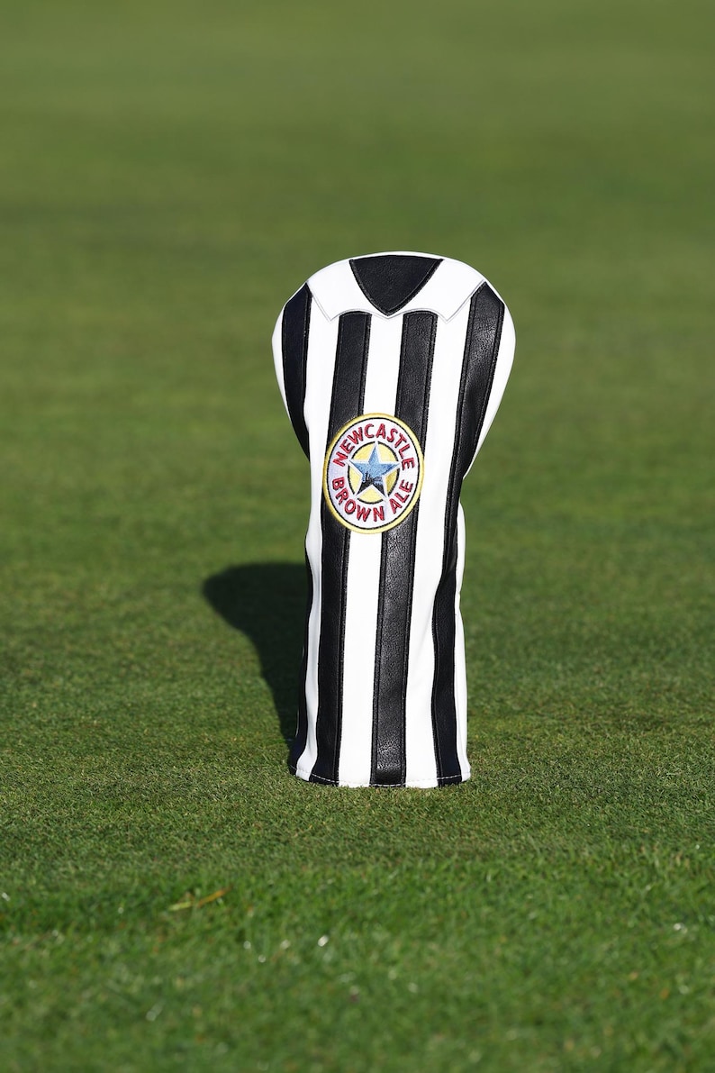 Peut inclure: Une housse de club de golf &agrave; rayures noires et blanches. La housse pr&eacute;sente un &eacute;cusson circulaire avec les mots "Newcastle Brown Ale" et une &eacute;toile. La housse est pos&eacute;e sur une surface herbeuse verte.