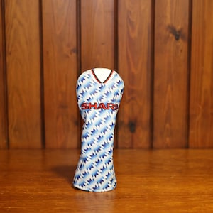 May include: A golf club head cover featuring a blue and white zigzag pattern, with the word "SHARP" in red. The cover has a V-neck design with red trim and a white collar. The background is a wooden surface.