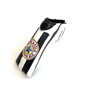 May include: A golf club head cover in black and white stripes. It has an embroidered Newcastle Brown Ale logo with a blue star and the text "ROWAY THE LADS".