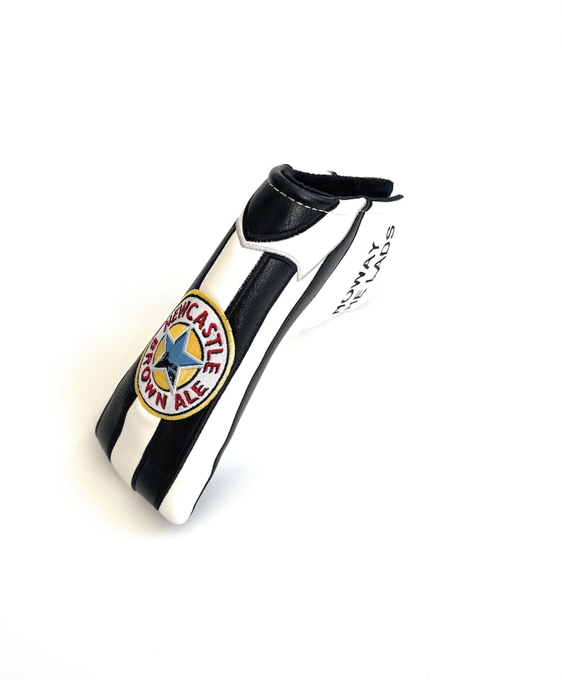 May include: A golf club head cover in black and white stripes. It has an embroidered Newcastle Brown Ale logo with a blue star and the text "ROWAY THE LADS".