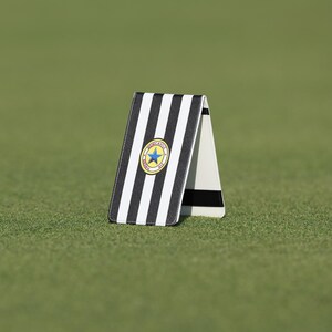 Può includere: Un segnaposto per pallina da golf con un design a strisce bianche e nere, con un emblema circolare con una stella blu e dettagli dorati. Il segnaposto è posizionato su una superficie erbosa verde.