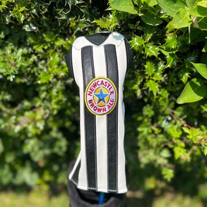 May include: A golf club head cover with a black and white striped design, resembling a sports jersey. The cover features a circular emblem with the text "Newcastle Brown Ale" and a blue star. The cover is in a golf bag.