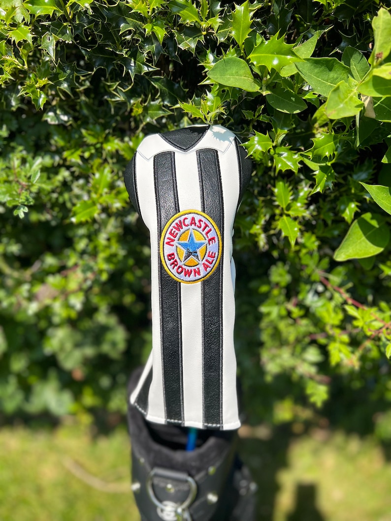 May include: A golf club head cover with a black and white striped design, resembling a sports jersey. The cover features a circular emblem with the text "Newcastle Brown Ale" and a blue star. The cover is in a golf bag.