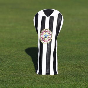 Peut inclure: Une housse de club de golf &agrave; rayures noires et blanches. La housse pr&eacute;sente un &eacute;cusson circulaire avec les mots "Newcastle Brown Ale" et une &eacute;toile. La housse est pos&eacute;e sur une surface herbeuse verte.