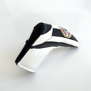 May include: A golf club head cover in white and black stripes, with a black interior. It features a circular emblem with the words "NEW BRAUNFELS" in yellow and blue. The cover is made from a leather-like material.