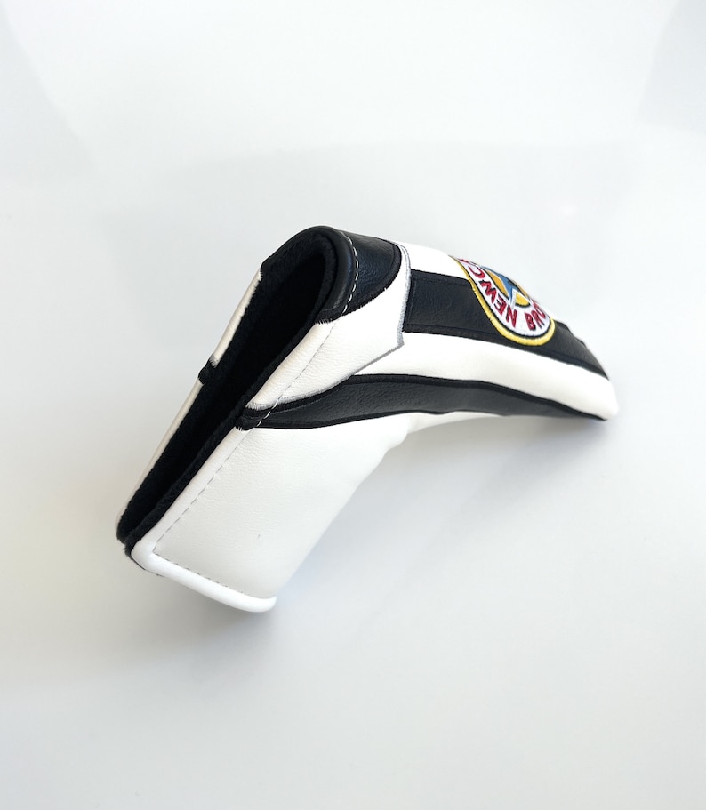 May include: A golf club head cover in white and black stripes, with a black interior. It features a circular emblem with the words "NEW BRAUNFELS" in yellow and blue. The cover is made from a leather-like material.