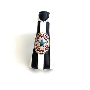 May include: A golf club head cover designed to resemble a Newcastle Brown Ale bottle. It features black and white stripes, a black top, and the Newcastle Brown Ale logo.