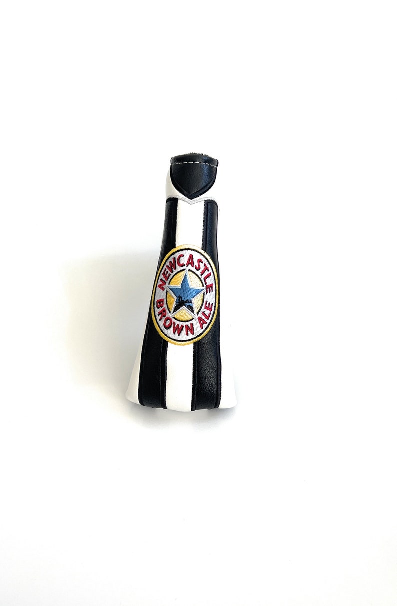 May include: A golf club head cover designed to resemble a Newcastle Brown Ale bottle. It features black and white stripes, a black top, and the Newcastle Brown Ale logo.