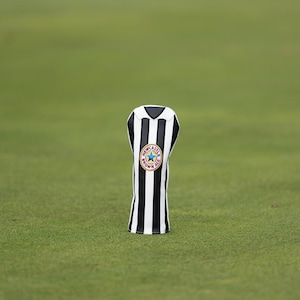 以下が含まれることがあります： 黒と白の縦縞模様のゴルフクラブヘッドカバー。カバーには、星と「Newcastle Brown Ale」の文字が入った円形のエンブレムが付いています。カバーは緑色の背景に配置されています。