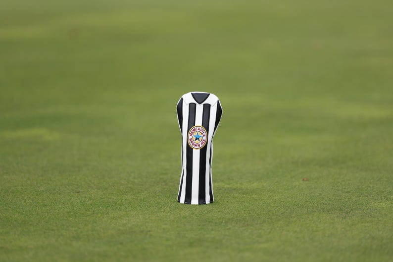 Peut inclure: Une housse de club de golf &agrave; rayures noires et blanches, orn&eacute;e d'un &eacute;cusson circulaire avec une &eacute;toile et l'inscription "Newcastle Brown Ale". L'ensemble est pos&eacute; sur un fond vert.