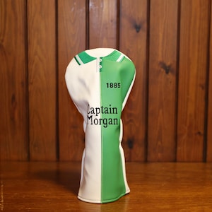 Pode incluir: Uma capa para a cabeça do taco de golfe em branco e verde. A capa é dividida verticalmente, com branco à esquerda e verde à direita. O texto "Captain Morgan" e o ano "1885" estão impressos no lado verde.