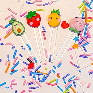 Puede incluir: Cinco adornos para pasteles con forma de fruta de colores con caras felices. Los adornos están hechos de fondant y están en palitos de plástico transparentes. Las frutas son una fresa, una piña, un melocotón, un aguacate y un corazón rojo.