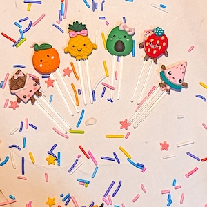 Puede incluir: Un conjunto de seis adornos coloridos en forma de comida de estilo de dibujos animados con caras felices. Los adornos están hechos de arcilla polimérica y están unidos a palitos delgados y blancos. Los adornos incluyen una piña, un aguacate, una fresa, una naranja, una sandía y una barra de chocolate. Los adornos están esparcidos sobre una superficie blanca con chispas de colores.
