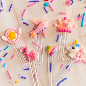 Peut inclure: Six pinces à cheveux en plastique colorées avec différents motifs, dont une licorne, un cœur, une étoile, un avion, un poney et une couronne. Les pinces sont sur un fond en bois clair avec des paillettes colorées.