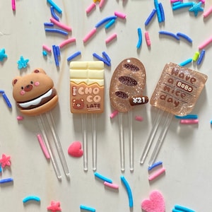 Peut inclure: Quatre marque-pages en résine colorés avec des motifs différents. Un ours brun portant un chapeau brun sur un hamburger, une barre de chocolat avec les mots "Choco Late" et "Bobo", une miche de pain brune avec le numéro "21", et un rectangle brun avec les mots "Have a nice day Bobo".