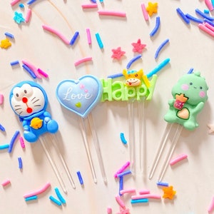 Peut inclure: Quatre décorations de gâteau colorées avec des bâtonnets en plastique transparent. Les décorations représentent un personnage de dessin animé bleu, un monstre de dessin animé vert, un cœur avec le mot "Love" et un mot "Happy" vert avec un animal de dessin animé jaune.