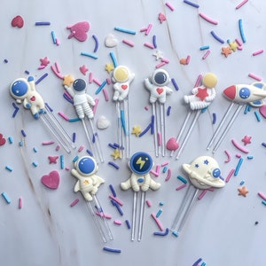 May include: A set of ten white astronaut-themed cake toppers with colorful details. The toppers are on clear plastic sticks and feature different designs, including astronauts, spaceships, and planets. The toppers are surrounded by colorful sprinkles.