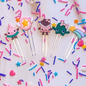 Peut inclure: Cinq trombones en papier de forme amusante et colorée avec des poignées en plastique transparent. Les personnages comprennent une pastèque, une étoile avec des ailes, une barre de chocolat, un cactus et un arc-en-ciel. Les trombones sont éparpillés sur une surface blanche avec des vermicelles roses, bleus et violets.