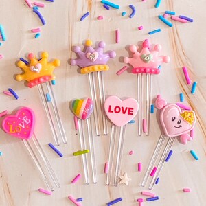 May include: Six colorful plastic bookmark clips with different designs.  The designs include a crown, a heart with the word "LOVE" on it, a rainbow heart, and a heart with a smiley face.  The clips are clear plastic and have a metal spring.