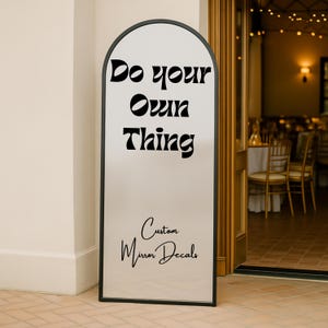 May include: A tall, arched mirror with a black frame. The mirror has the words "Do your Own Thing" in black script at the top, and "Custom Mirror Decals" at the bottom. The mirror is standing in front of a doorway.